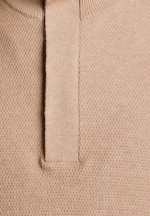 Beżowy sweter z teksturowanym wzorem i krótkim kołnierzykiem. Tkanina ma miękkie, dziane wykończenie z wzorem w kształcie diamentu.