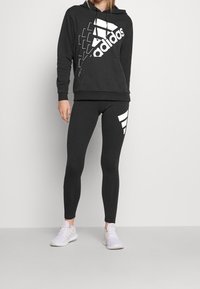 Sweat à capuche noir avec grand logo "adidas" blanc et motif géométrique, assorti à un legging noir avec des accents de bandes blanches. Baskets blanches.