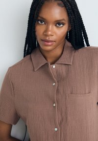 Chemise marron à boutons avec un tissu texturé. Manches courtes, col, et une poche sur le côté gauche, avec des boutons clairs.