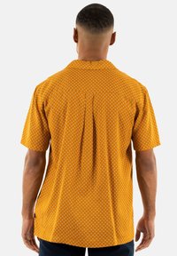 Korte mouwen shirt in mosterdgeel met een geometrisch wit patroon. Voorzien van een spread collar en een rugplooitje voor gemakkelijke beweging.