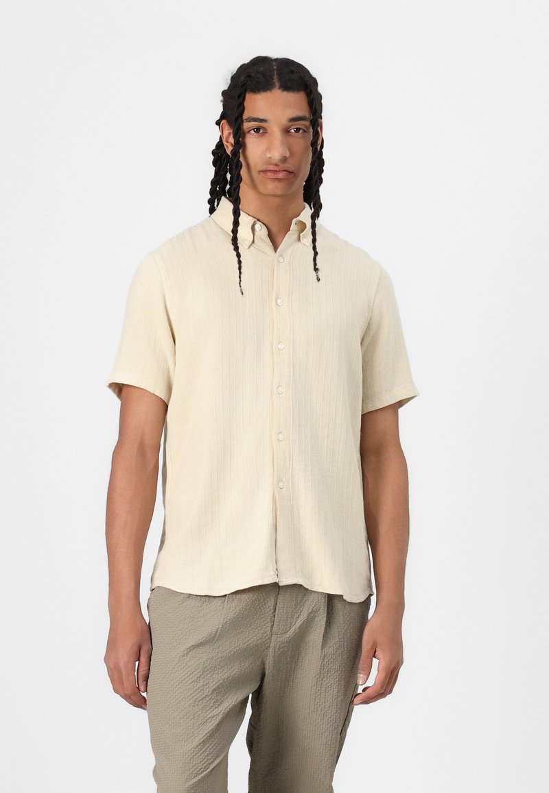 Korte mouwen ribbelshirt in lichtbeige, met een button-down kraag en knopen aan de voorkant. Draagt bij gebreide lichtgrijze broek.