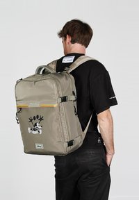 Mochila color caqui con un acabado brillante, que presenta un gráfico de Mickey Mouse, una franja amarilla de acento, correas ajustables y múltiples compartimentos.