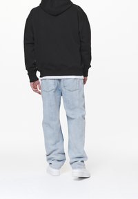 Felpa nera con cappuccio, jeans di denim azzurro chiaro e sneakers bianche. Cuciture visibili e vestibilità rilassata, con una maglietta bianca sotto.