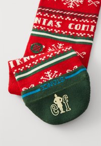 Calcetines rojos con un diseño de copos de nieve blancos, rayas verdes y blancas. Incluyen un talón verde y acentos azules, con un logo de "elfo" en color crema.