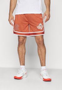 Pantalones cortos de baloncesto de malla naranja con acentos blancos, que presentan un logo en el muslo izquierdo y un parche gráfico en la parte delantera derecha.