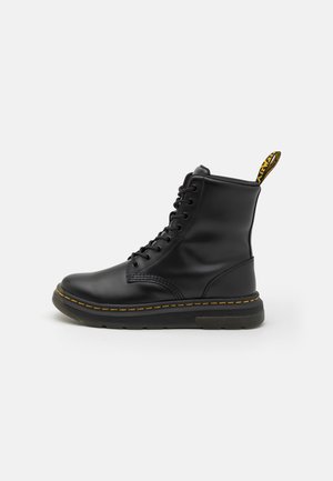 Bottine à lacets en cuir noir avec un accent jaune sur la languette, un col rembourré et une semelle épaisse noire avec un détail de couture jaune.