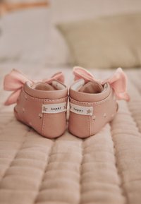 Paire de petites chaussures de bébé roses avec des nœuds et des étiquettes "happy" à l'arrière, placées sur une surface matelassée douce.