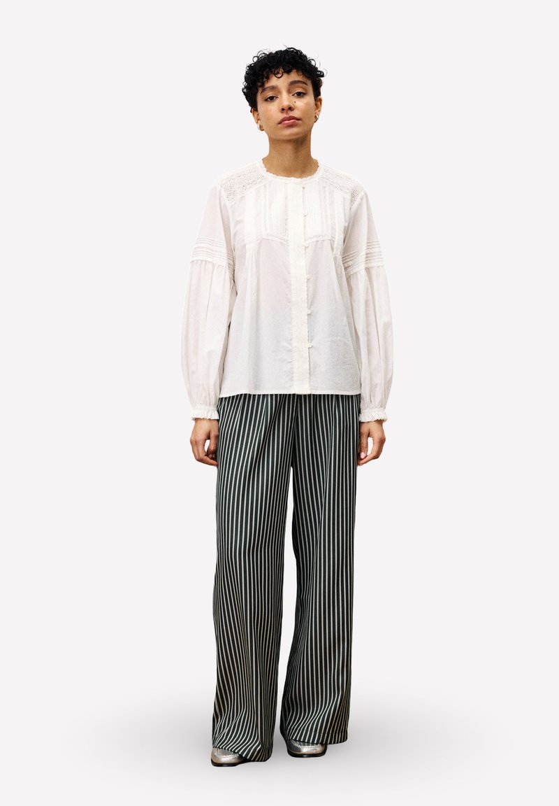 Blouse blanche avec des détails texturés, bouton à l'avant et manches longues. Associée à un pantalon large à rayures noir et blanc et des chaussures métalliques.