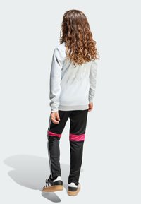 Joven con cabello rizado y largo, vistiendo sudadera blanca, pantalones deportivos negros con franjas rosas en las rodillas y zapatillas negras, de espaldas a la cámara.