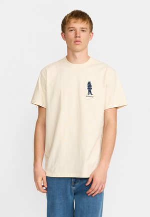Joven con cabello claro que lleva una camiseta crema con un pequeño gráfico azul marino de una figura que lleva platos y la palabra "Revolución" en el pecho.