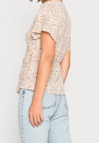 Mujer vistiendo una blusa corta con mangas de flores en tono rosa claro, combinada con unos jeans de tiro alto en azul claro, mostrada de lado.