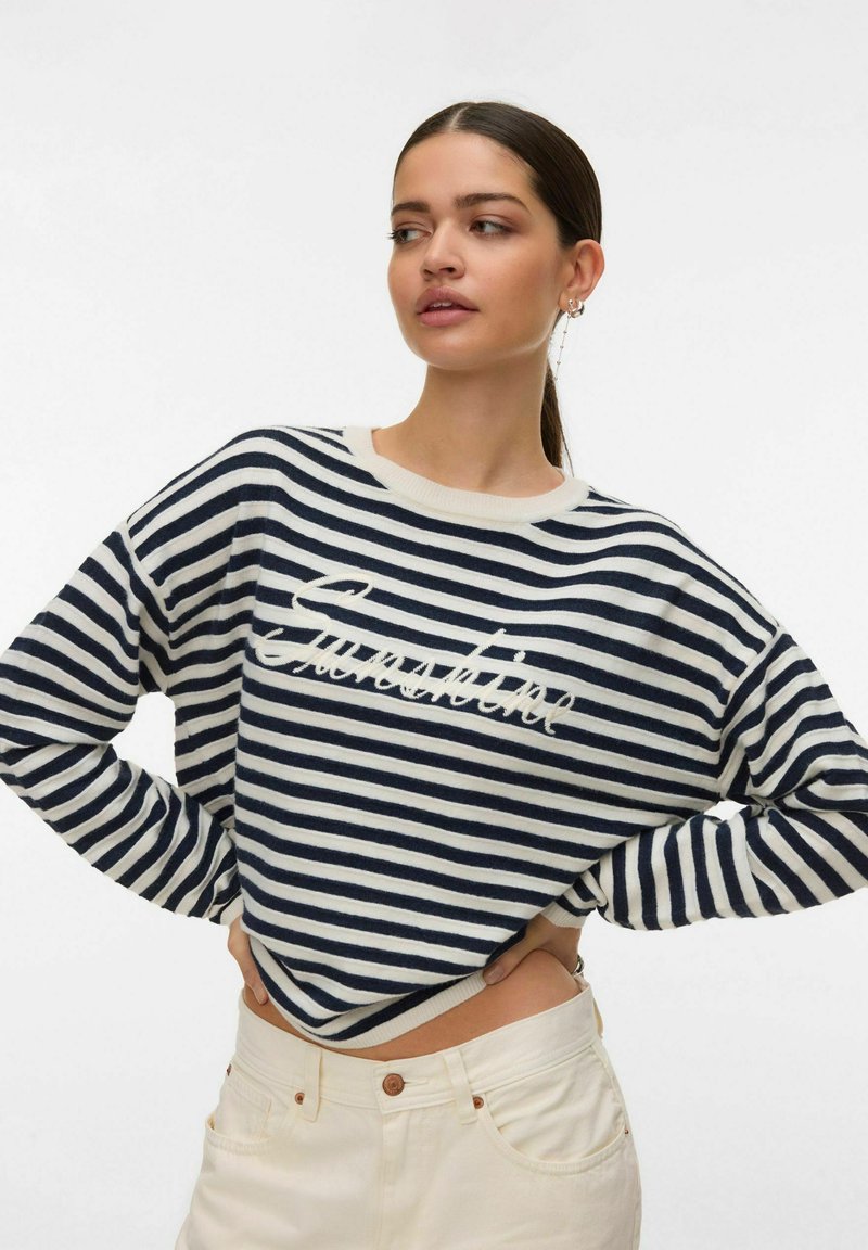 Donna con capelli scuri raccolti in una coda di cavallo, che indossa un maglione a righe blu navy e bianche con testo in corsivo, abbinato a pantaloni a vita alta color avorio, con le mani sui fianchi.