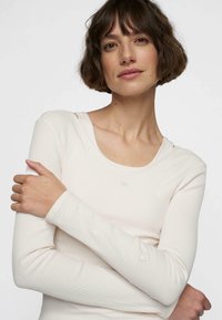 Top lungo a maniche di colore beige chiaro in morbido tessuto, con scollo rotondo e design aderente. Presenta un delicato dettaglio del logo sulla manica.