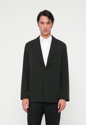 Schwarze Blazer aus leichtem Stoff, taillierte Passform, Ein-Knopf-Verschluss, zwei Vordertaschen und breite Revers, getragen über einem weißen Hemd.