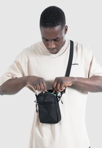 Zwarte nylon schoudertas met verstelbare riem, voorzien van een vak met rits en trekkoordsluiting, vastgehouden door een persoon die een beige shirt draagt.