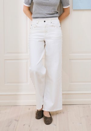 Személy magasderekú, széles szárú, oldalt szegecselt fehér nadrágban, szürke rövid ujjú felsőben és barna balerinacipőben, fapadlón állva.