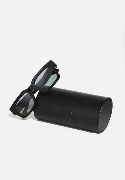 Black rectangular sunglasses with tinted lenses and thick frames, placed next to a matte black cylindrical case featuring embossed logo text.