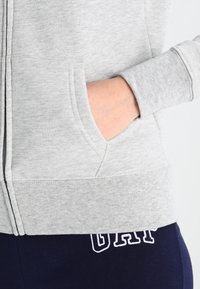 Sweat à capuche gris zippé avec une poche kangourou, des poignets et une taille côtelés, associé à un pantalon bleu marine avec des lettres blanches brodées.