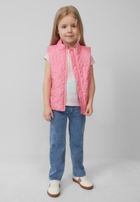 Roze gewatteerde mouwloze vest met een hoge kraag, gedragen over een licht shirt, gecombineerd met spijkerbroeken en witte sneakers met bruine accenten.