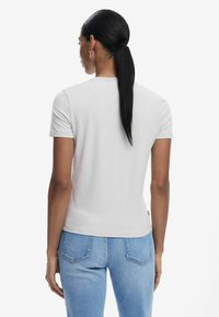 T-shirt grigio chiaro con maniche corte e design aderente. Il tessuto appare liscio. Indossato con jeans blu, mette in mostra uno stile semplice e casual.