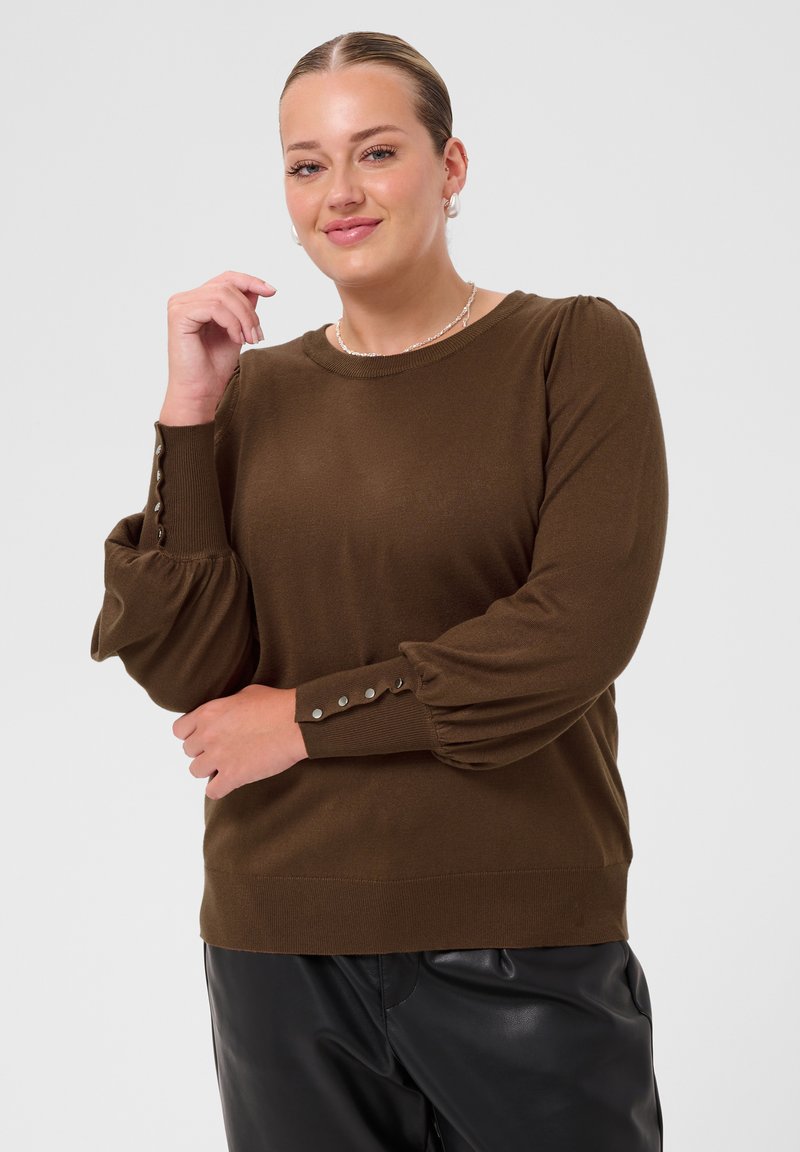 Femme aux cheveux plaqués en arrière portant un pull marron à manches longues avec des poignets boutonnés et un pantalon en cuir noir, debout devant un fond uni.