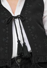Zwarte vest met geborduurde bloemenpatronen, met een voorknoop en franjes. Draag boven een wit shirt, gecombineerd met donkere broek.