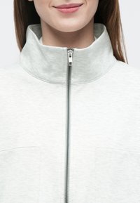 Lysegrå genser med høy krage og glidelås foran. Har en glatt tekstur og en brystlomme på venstre side.