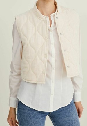 Pehmeä toppaliivi pyöreällä kaula-aukollla, napeilla ja aaltoilevalla kuviolla, valkoisen napillisen paidan päällä. Pehmeä rakenne, lyhyt malli.