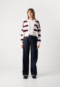 Cardigan a righe in crema, rosso e blu navy, con texture a coste, scollatura a V profonda e bottoni dorati, abbinato a jeans larghi in denim scuro.