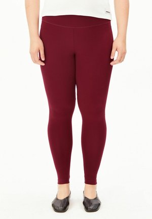 Personne portant des leggings rouge foncé et des chaussures plates noires, debout devant un fond blanc.