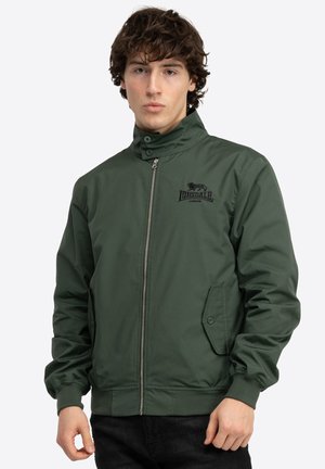 Giacca bomber verde con zip frontale, colletto alto, polsini elasticizzati e tasche laterali. Presenta un logo sul petto. Tessuto dalla consistenza liscia.