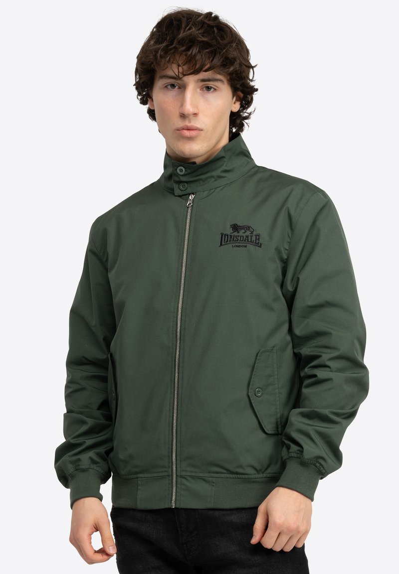 Chaqueta bomber verde con cremallera frontal, cuello alto, puños elásticos y bolsillos laterales. Presenta un logo en el pecho. Textura de tela suave.