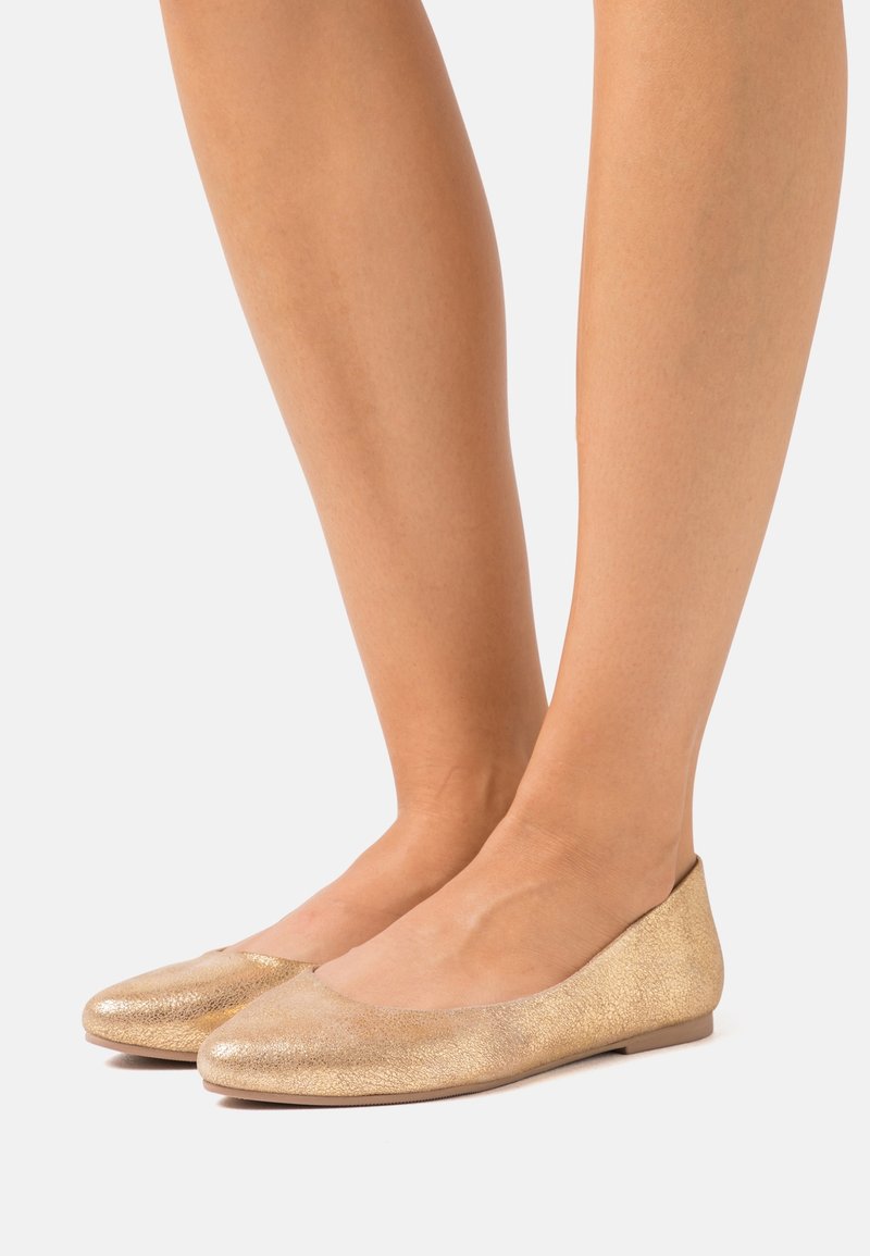 Jambes portant des chaussons de ballet plats dorés métalliques avec un embout arrondi sur un fond clair uni.