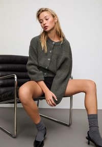 Pletená zelená kardigán s oversized rukávmi a šedými gombíkmi, nosená cez čierne tričko. Model sedí na chrómovej stoličke, v kombinácii so šedými ponožkami a lodičkami.