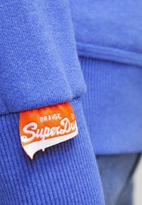 Tejido de punto azul con un acabado texturizado, con una etiqueta naranja que dice "SuperDry" en blanco con "ORANGE" encima.