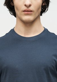 Νεαρός ενήλικας που φοράει ένα απλό σκούρο μπλε μπλουζάκι με στρογγυλή λαιμόκοψη, εμφανιζόμενος από τους ώμους μέχρι τα χείλη σε ανοιχτό φόντο.