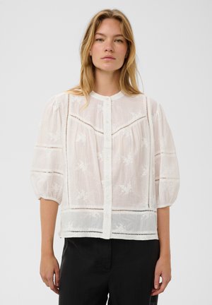 Vrouw draagt een witte doorschijnende blouse met bloemrijke borduursels, knopen aan de voorkant en driekwart pofmouwen, gecombineerd met een zwarte broek.