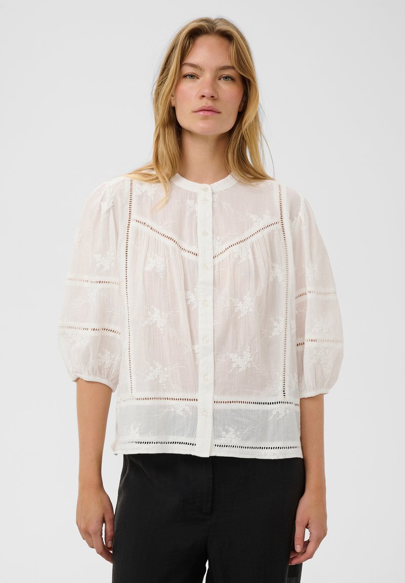 Femme portant un chemisier blanc transparent avec broderies florales, fermeture à boutons devant et manches ballon trois-quarts, associé à un pantalon noir.