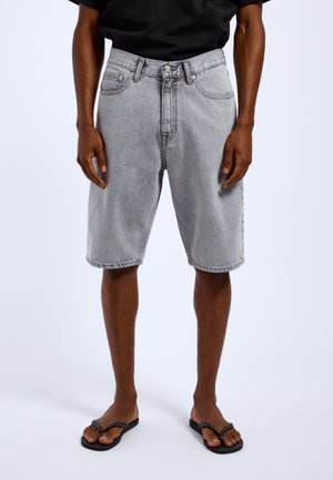 Mand iført lysegrå denimshorts, sort t-shirt og sorte flip-flops står på en hvid baggrund.
