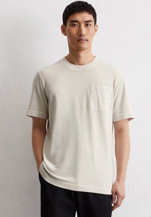 Lichtbeige T-shirt met korte mouwen, ronde hals en een klein borstzakje. Zachte textuur en een comfortabele pasvorm. G gedragen met zwarte broeken.