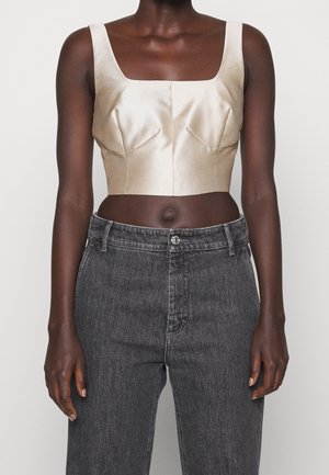Osoba o ciemnej karnacji ubrana w kremowy, satynowy, krótki top na ramiączkach i wysokiej talii wyblakłe czarne dżinsy, stojąca na tle jednolitego białego tła.