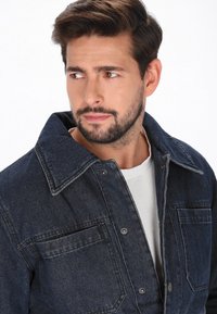 Sötét farmerdzseki nagy gallérral, két felső zsebbel és ezüst színű fém részekkel. Az anyag finom textúrájú, kontrasztos varrási részletekkel.