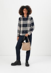 Langärmliges kariertes Hemd in Grau und Marineblau, mit Knopfleiste, kombiniert mit Marineblauen Hosen und schwarzen klobigen Stiefeln, hält eine Handtasche aus beigem Wildleder.