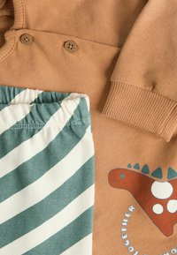 Ruskea huppari napituksilla ja dinosaurusteemalla, yhdistettynä vihreisiin ja valkoisiinraitapolkimiin, joissa on joustovyötärö. Puuvillakangasta, rento muotoilu.