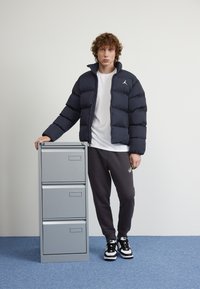 Puhasta jakna v temno modri barvi, bela majica, sivi trenirki, stoji ob sivi omari za dokumente s tremi predali, na modrem preprogi.