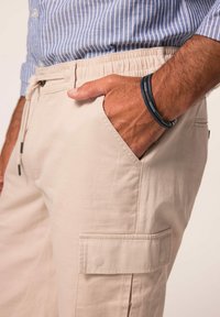 Beige cargo bukser med elastisk talje, sidelommer og en struktureret stof. Modellen har hånden i lommen og bærer et sort armbånd.