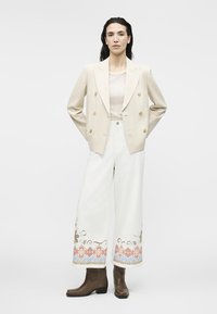 Γυναίκα όρθια, φοράει μπεζ σακάκι διπλής σειράς κουμπιών, λευκό παντελόνι με φαρδιά μπατζάκια και πολύχρωμη κεντητά στο τελείωμα, καθώς και καφέ μποτάκια αστραγάλου.