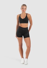 Zwarte sportbeha met een V-hals en geplooide details, gecombineerd met zwarte shorts met hoge taille. Beide zijn gemaakt van een glad, rekbaar materiaal.