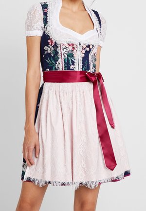 Kobieta ubrana w kwiecistą sukienkę dirndl z białymi koronkowymi rękawami, różowym koronkowym fartuchem oraz szeroką ciemnoczerwoną satynową kokardą zawiązaną w talii.