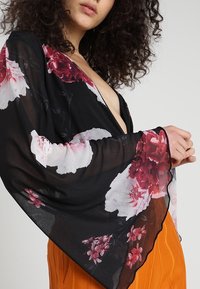 Kimono nero trasparente con grandi motivi floreali nelle tonalità di rosso e rosa; presenta maniche ampie e fluide e un bordo in pizzo.